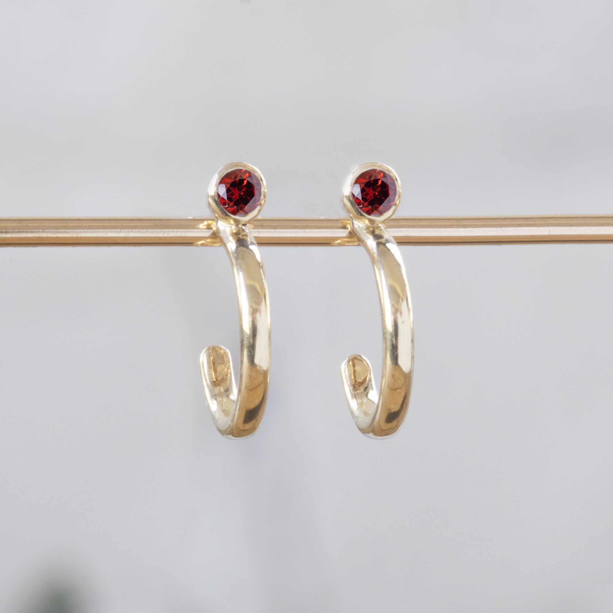 Custom Garnet Birthstone Hoop Earrings | 14K Yellow Gold. Displayed on holding bar with white blurred background. 