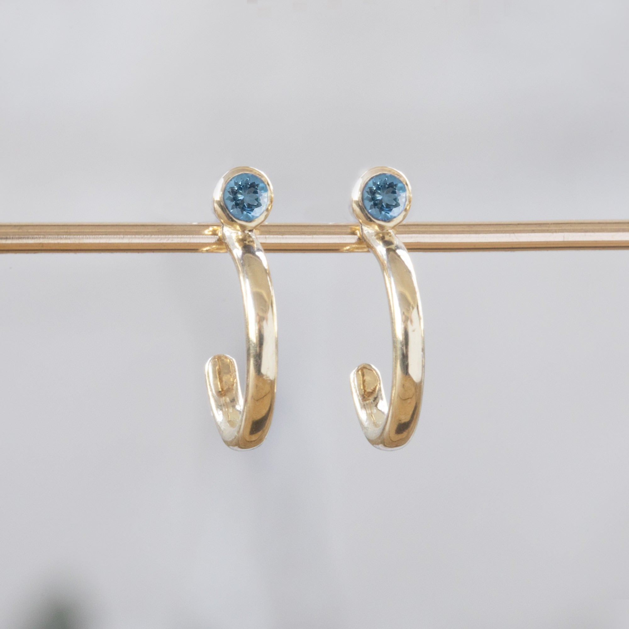 Custom Aquamarine Birthstone Hoop Earrings | 14K Yellow Gold displayed on bar with white blurred background. 