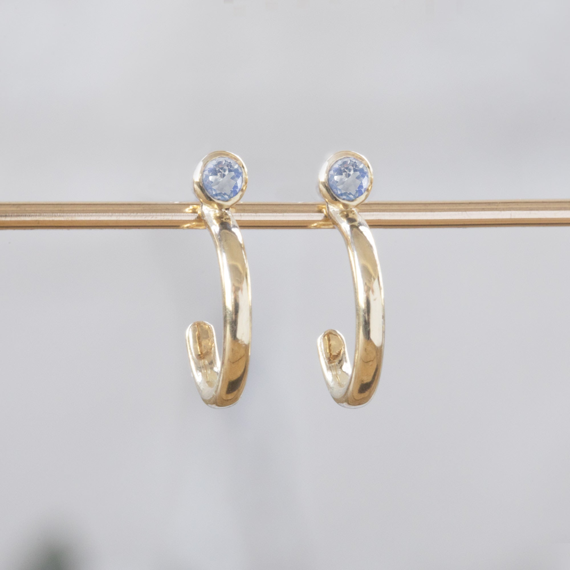 Custom Moonstone Birthstone Hoop Earrings | 14K Yellow Gold. Displayed on holding bar with white blurred background. 
