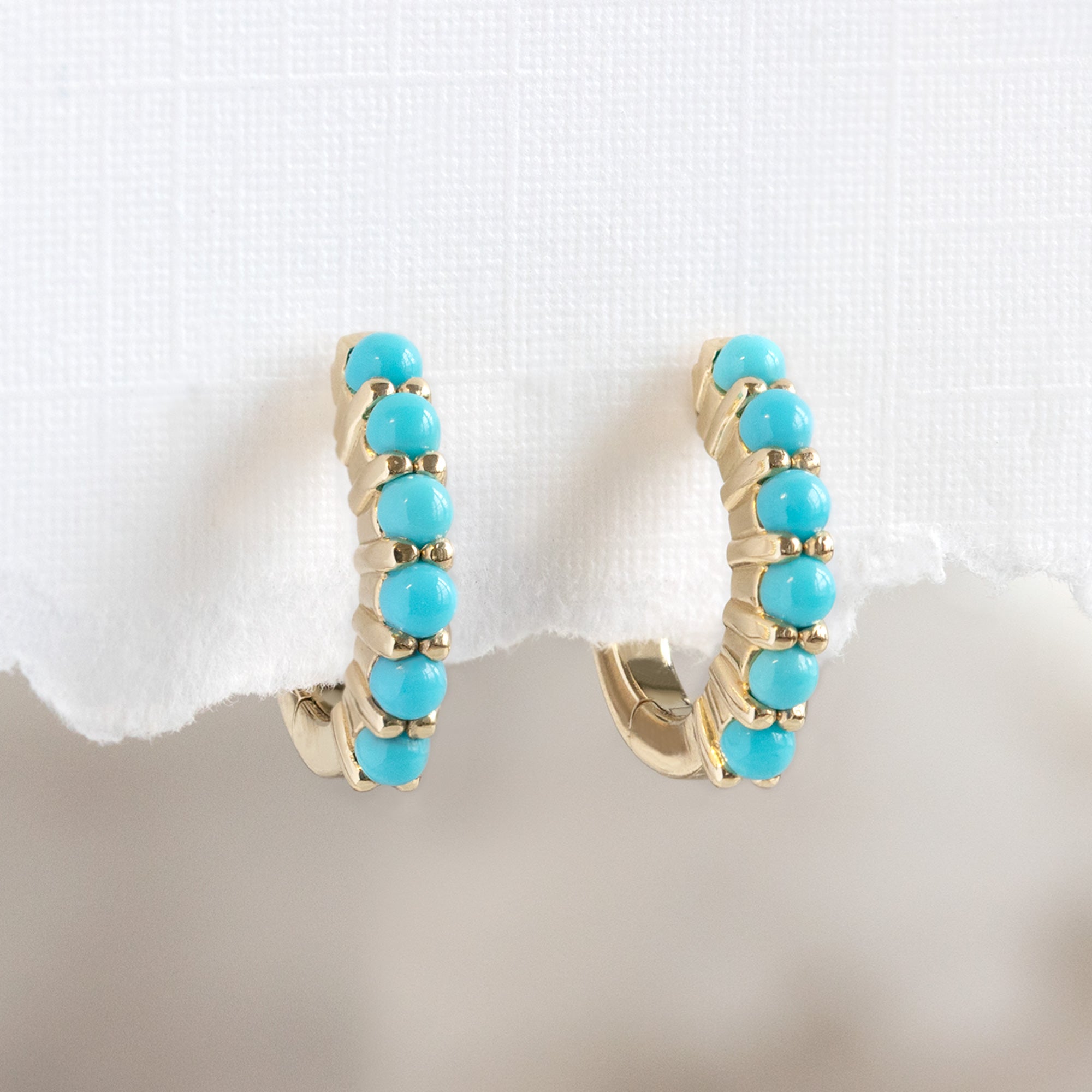 The Turquoise Huggie Hoop Earrings in 14K Yellow Gold hanging on white textured paper