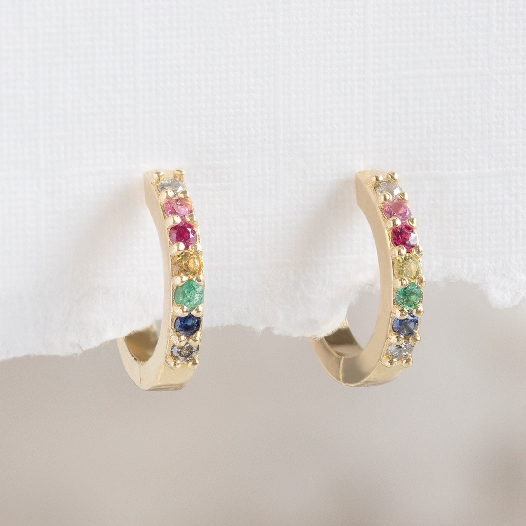 The Rainbow Sapphire Pavé Huggie Hoops in 14K Yellow Gold hanging on white textured paper