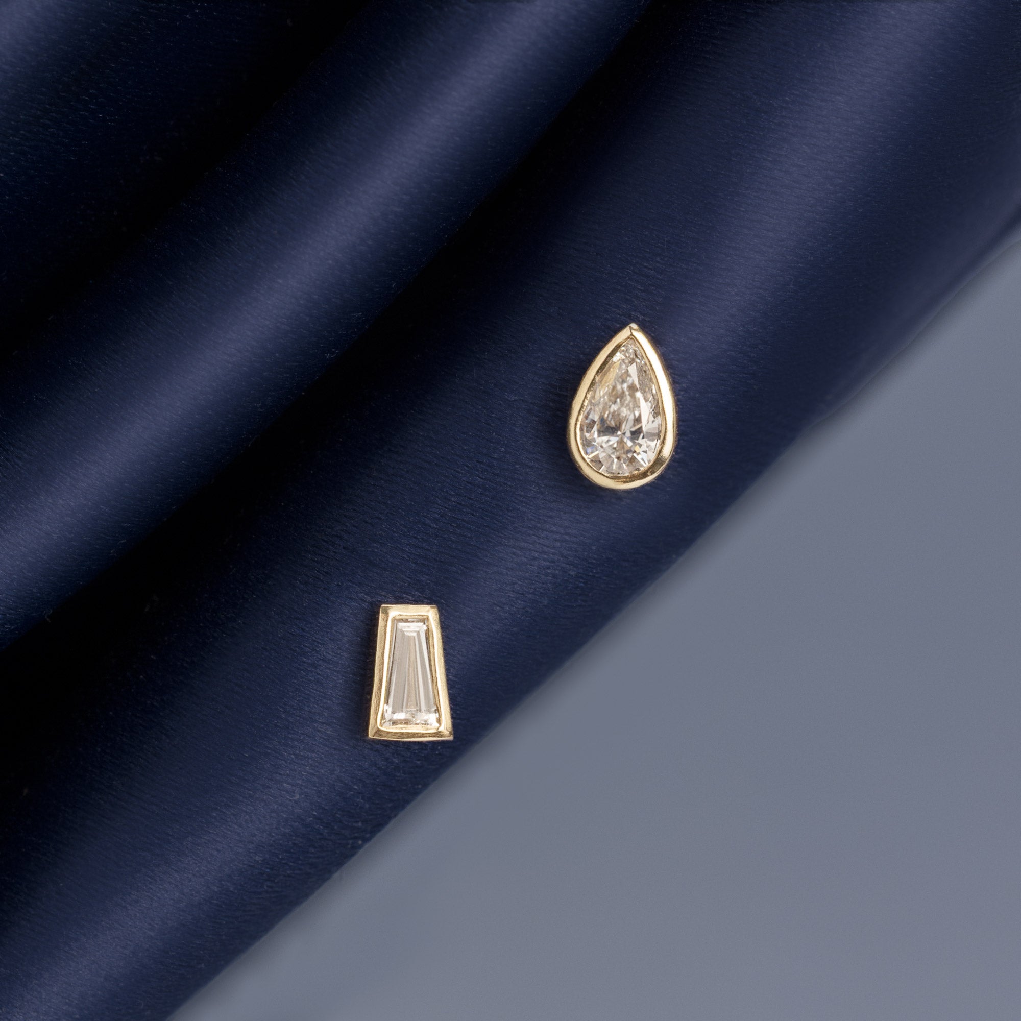 ‘It Girl' Baguette + Pear Diamond Stud Earrings in 14K Yellow Gold hanging on navy blue fabric