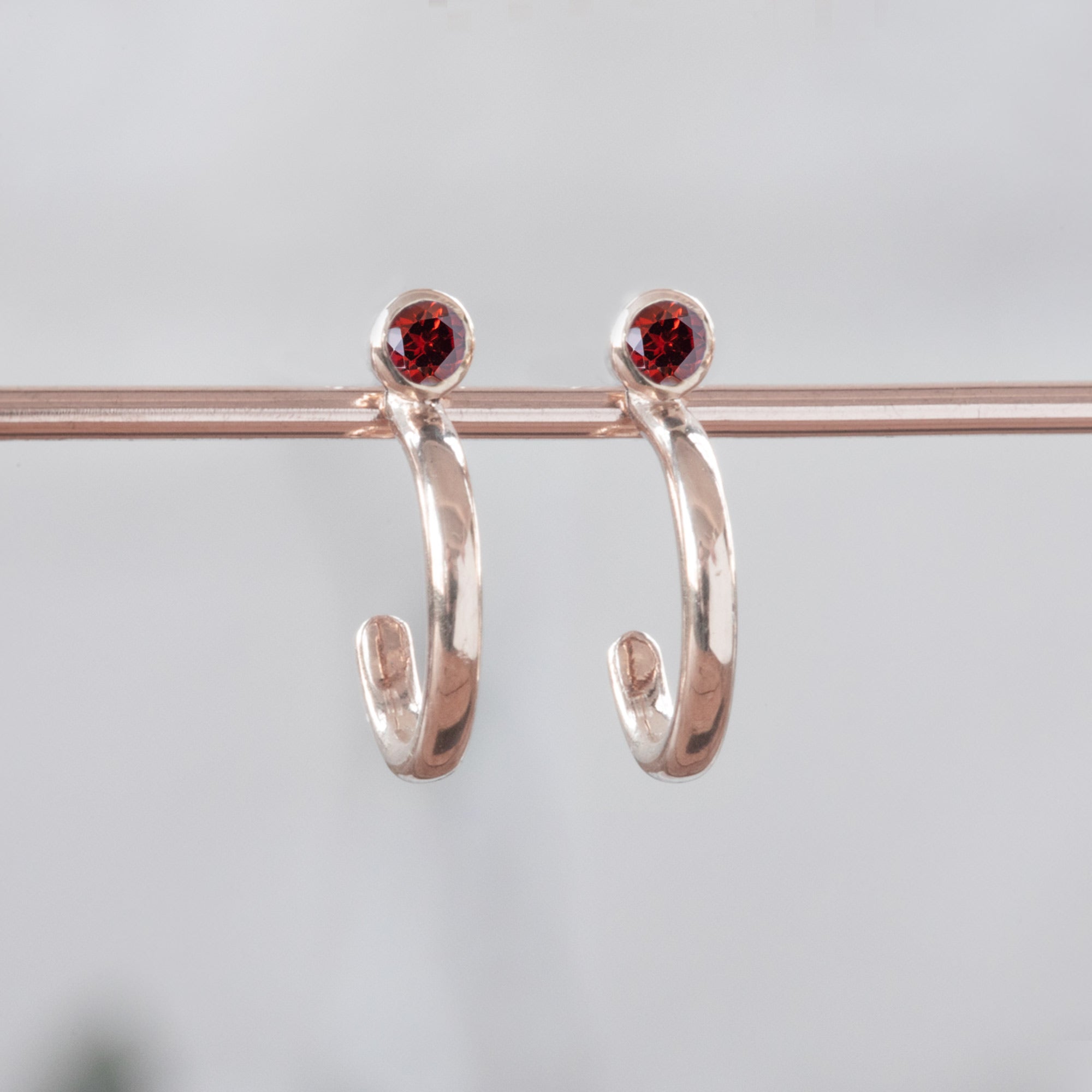 Custom Garnet Birthstone Hoop Earrings | 14K Rose Gold. Displayed on holding bar with white blurred background. 