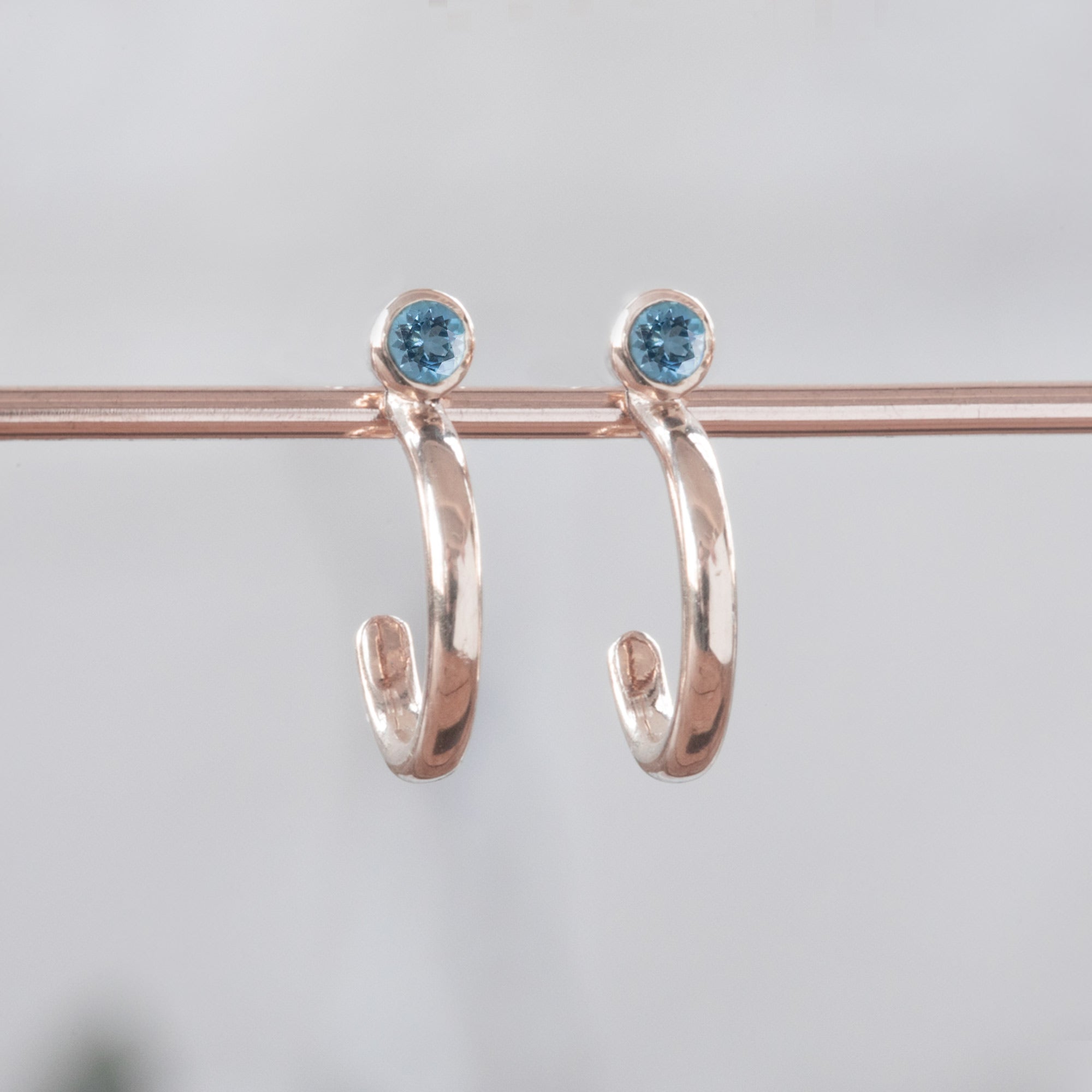 Custom Aquamarine Birthstone Hoop Earrings | 14K Rose Gold displayed on bar with white blurred background. 