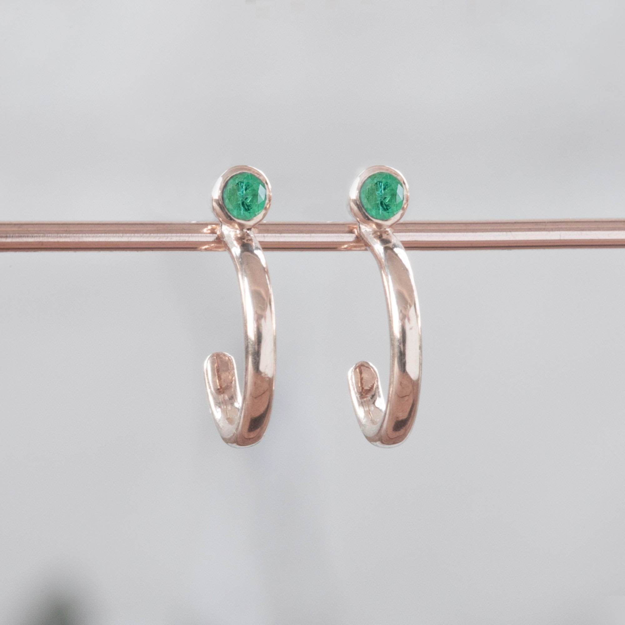 Custom Emerald Birthstone Hoop Earrings | 14K Rose Gold displayed on holding bar with white blurred background. 