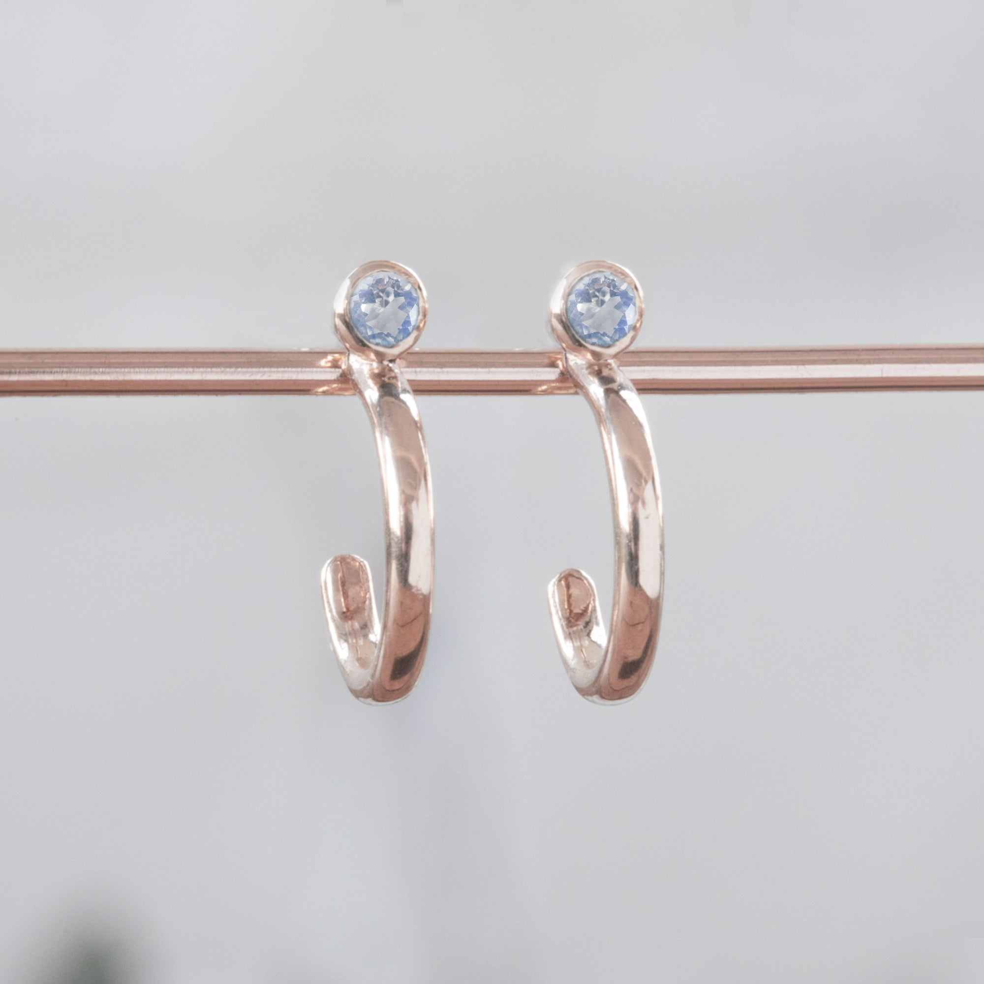 Custom Moonstone Birthstone Hoop Earrings | 14K Rose Gold. Displayed on holding bar with white blurred background. 