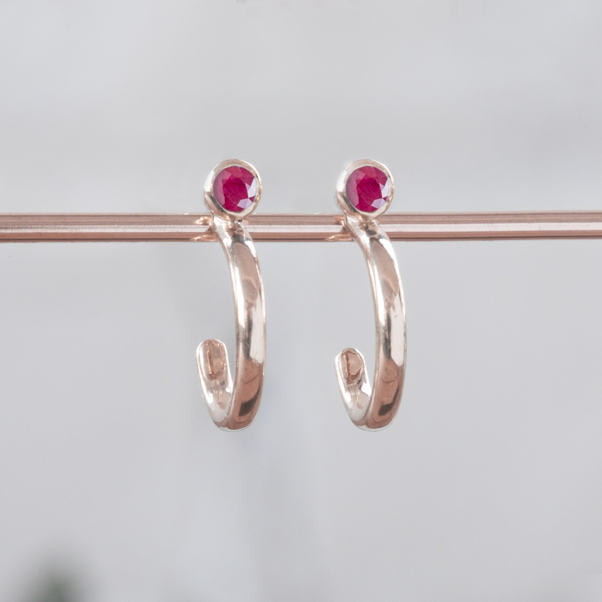 Custom Ruby Birthstone Hoop Earrings | 14K Rose Gold. Displayed on holding bar with white blurred background. 