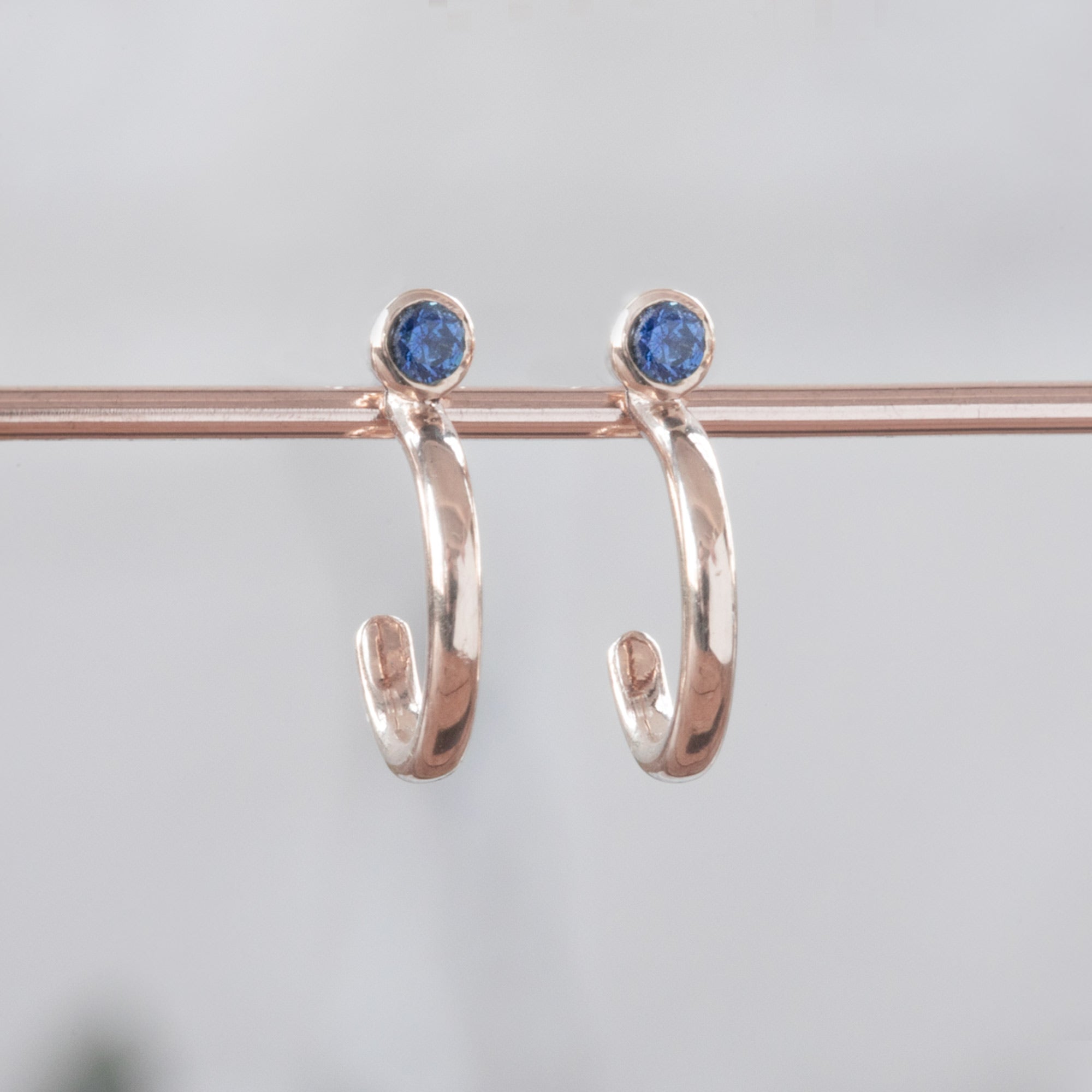 Custom Blue Sapphire Birthstone Hoop Earrings | 14K Rose Gold displayed on bar with white blurred background. 