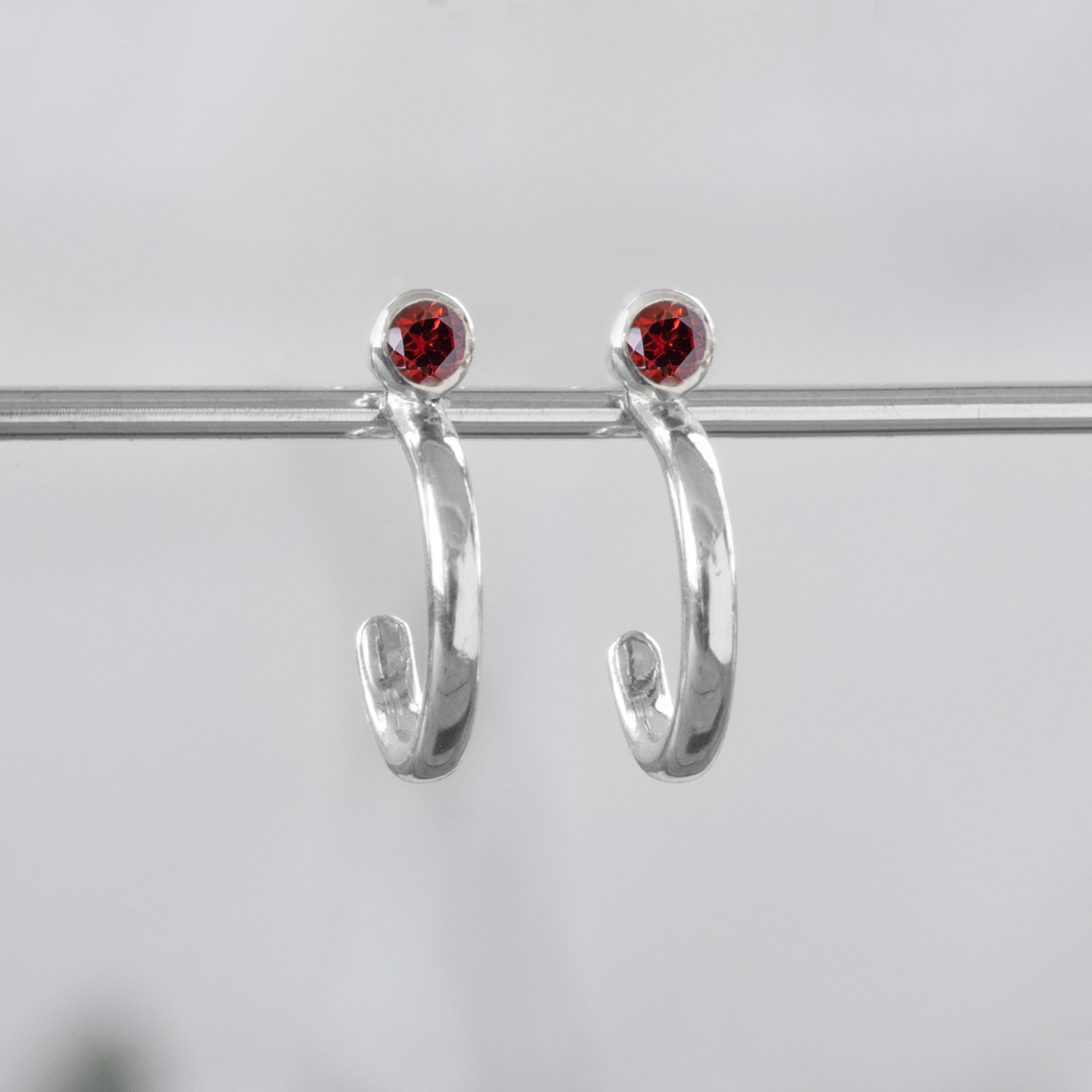 Custom Garnet Birthstone Hoop Earrings | 14K White Gold. Displayed on holding bar with white blurred background. 