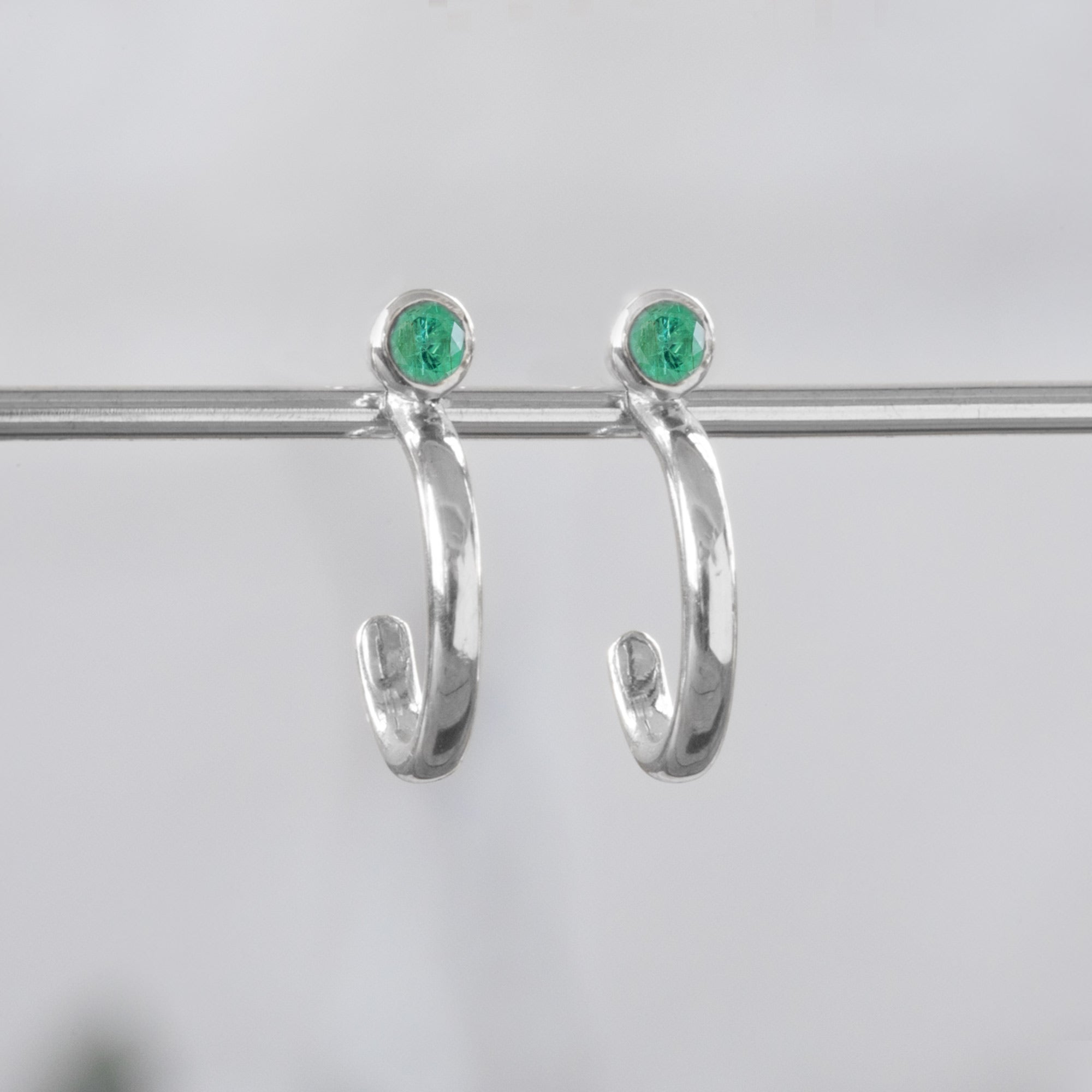 Custom Emerald Birthstone Hoop Earrings | 14K White Gold displayed on holding bar with white blurred background. 