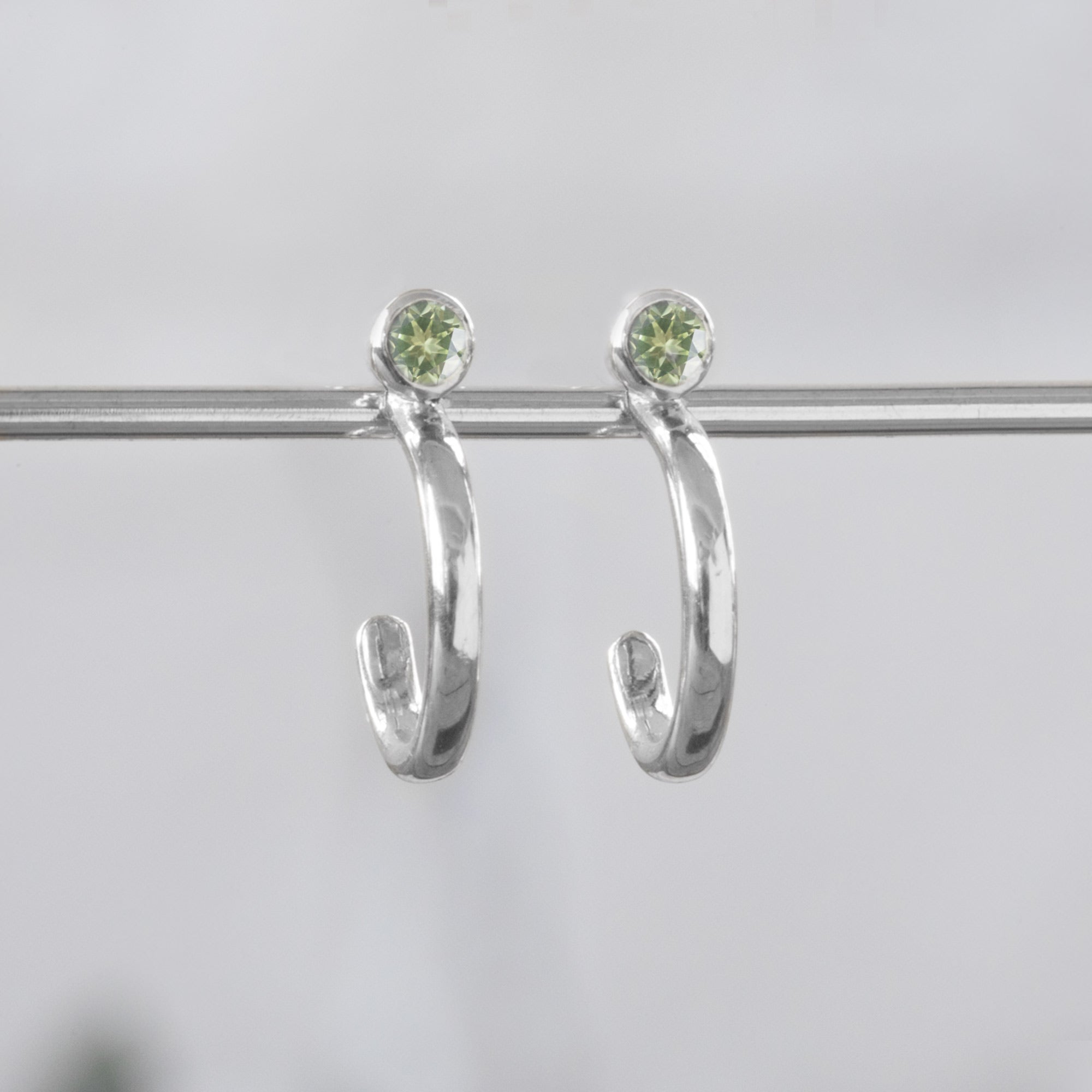 Custom Peridot Birthstone Hoop Earrings | 14K White Gold. Displayed on holding bar with white blurred background. 