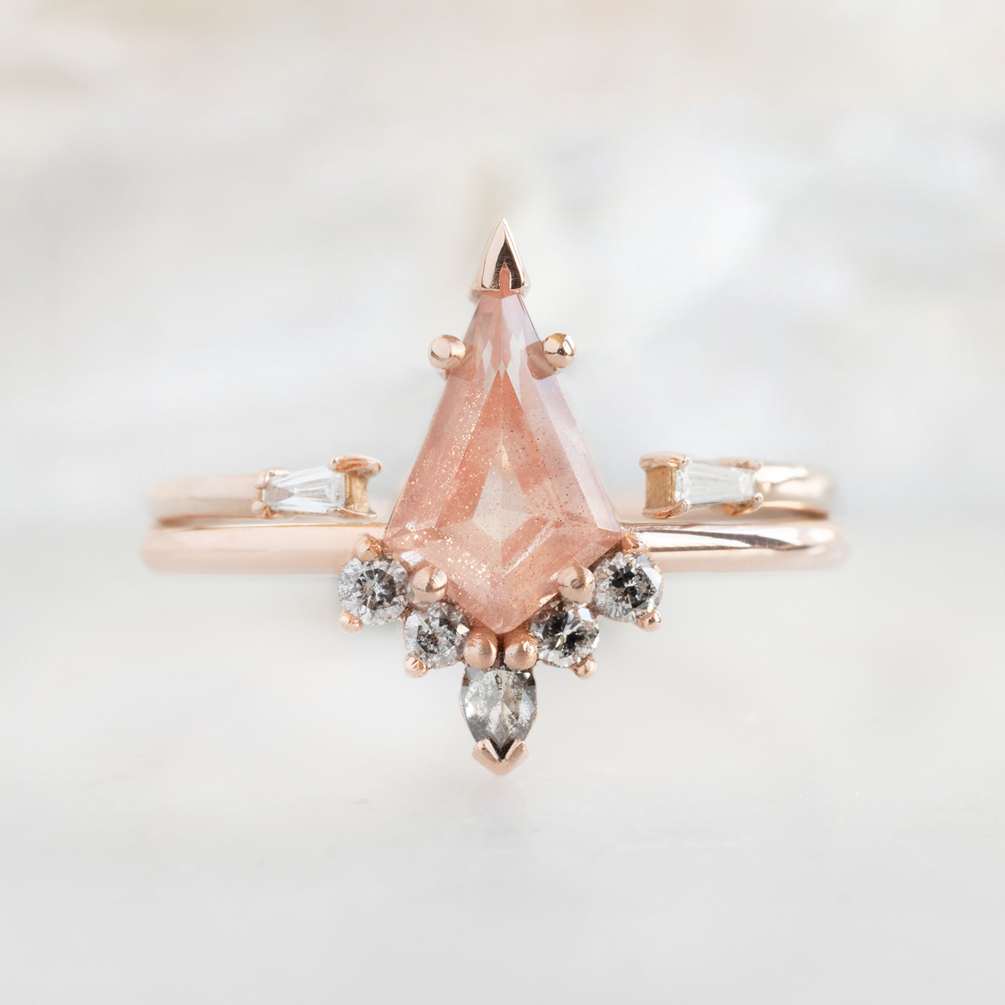 The Aster Ring | 0.79 carat Kite Sunstone in 14K Rose Gold with white blurred background. Displayed next to the open cuff white diamond baguette wedding band. 