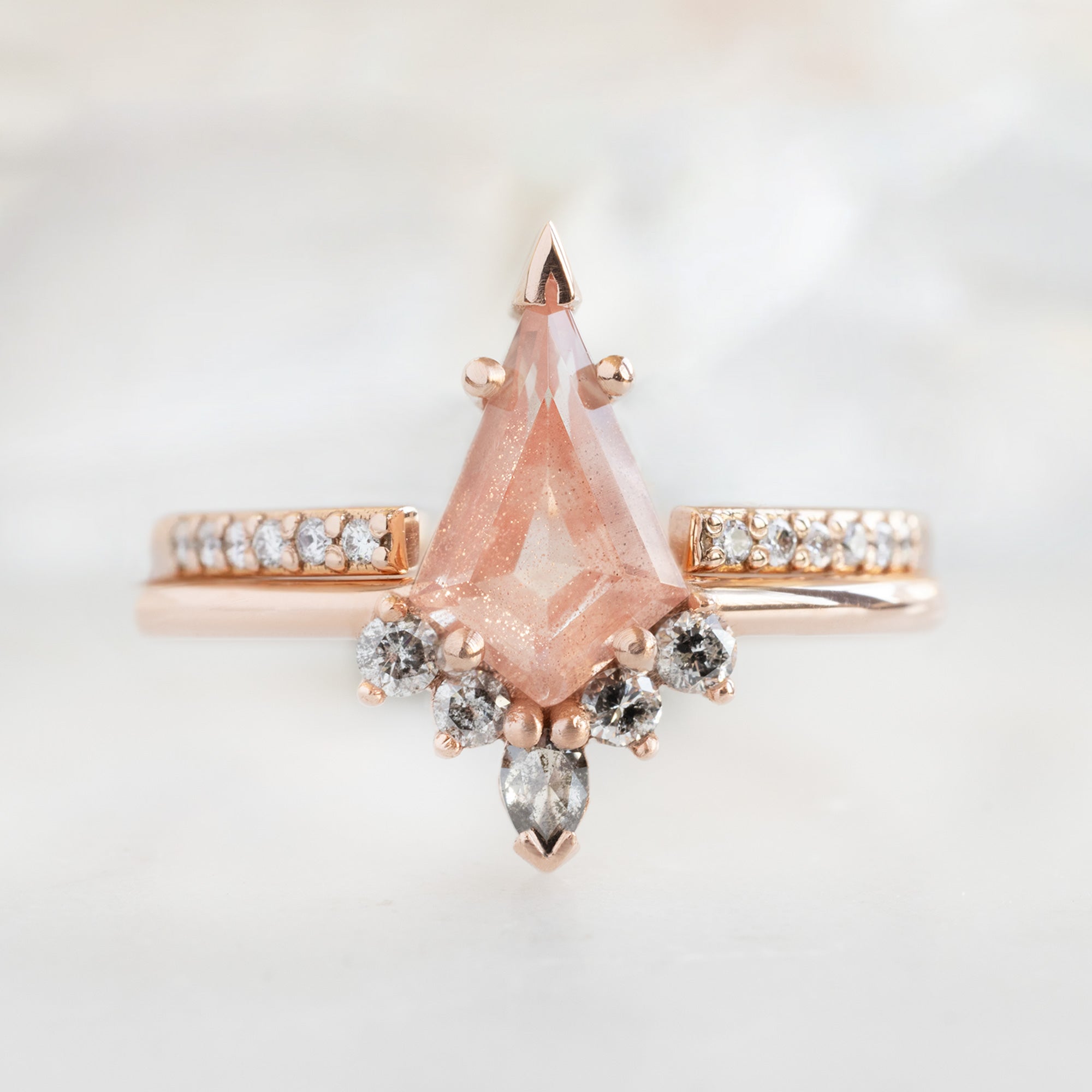 The Aster Ring | 0.79 carat Kite Sunstone in 14K Rose Gold with white blurred background. Displayed next to the open cuff white diamond pavé wedding band. 