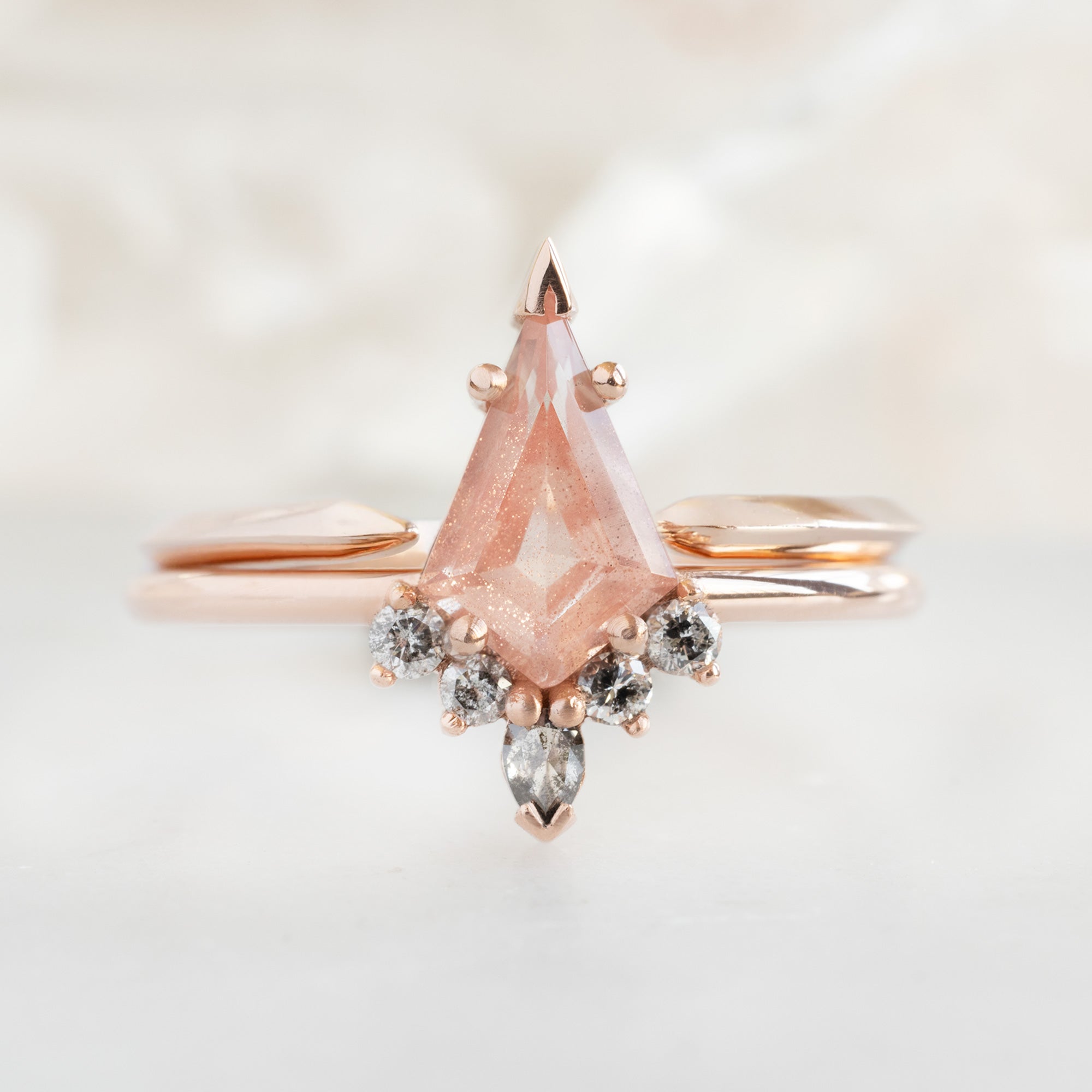 The Aster Ring | 0.79 carat Kite Sunstone in 14K Rose Gold with white blurred background. Displayed next to the open cuff knife edge wedding band. 