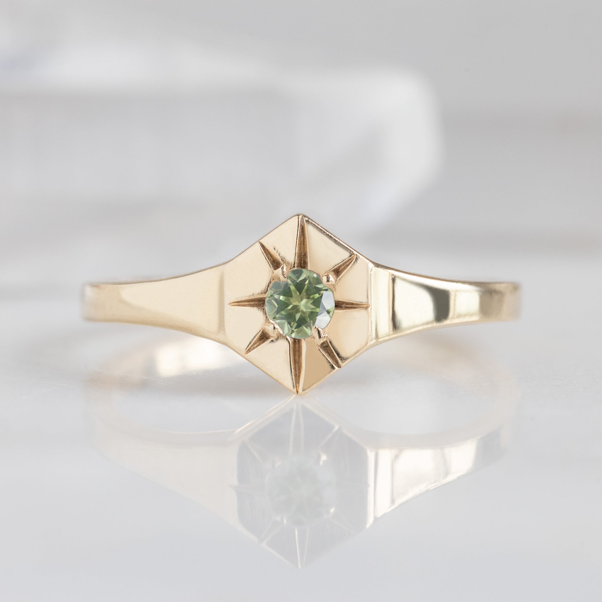 Custom Peridot Birthstone Signet Ring | 10K Yellow Gold. Displayed on marble surface with white blurred background. 