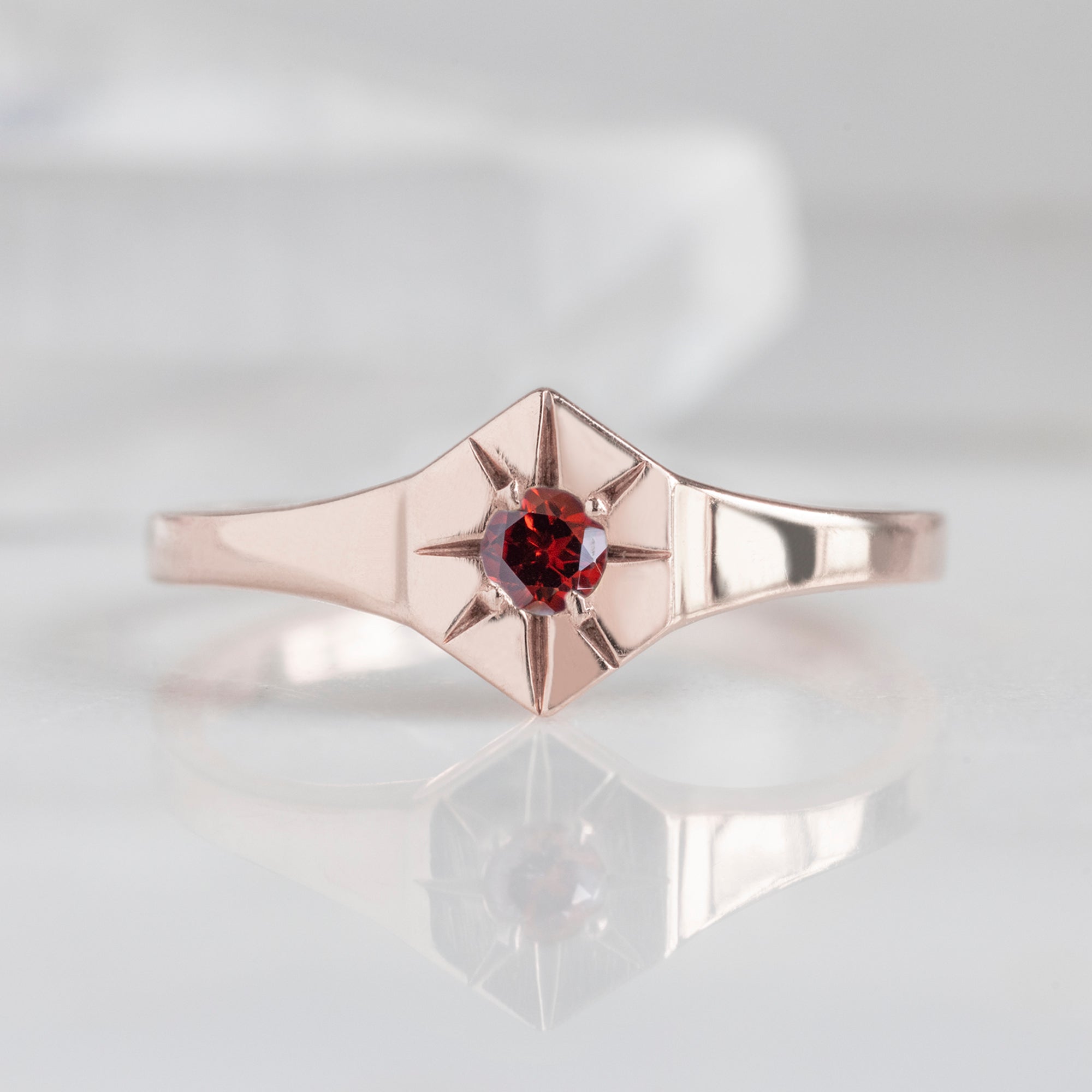 Custom Garnet Birthstone Signet Ring | 10K Rose Gold. Displayed on marble surface with white blurred background. 