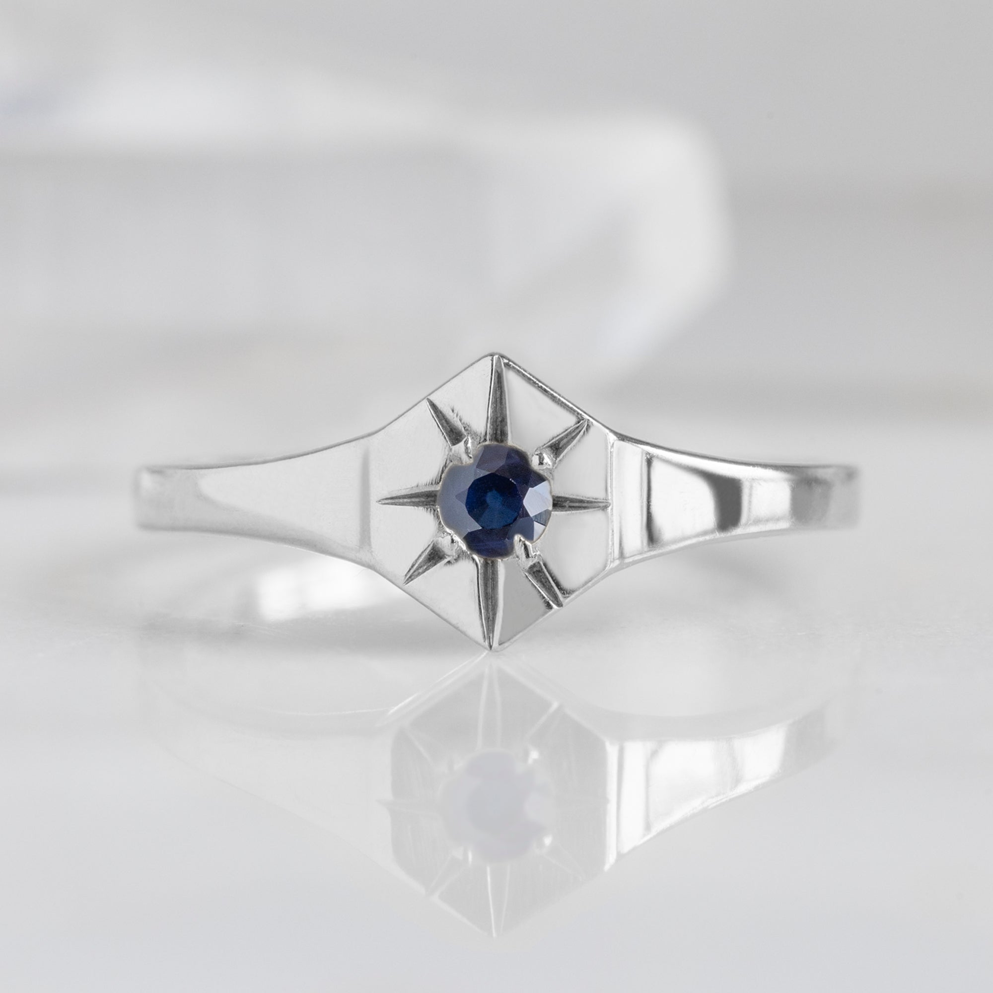 Custom Blue Sapphire Birthstone Signet Ring | 10K White Gold displayed on marble surface with white blurred background.
