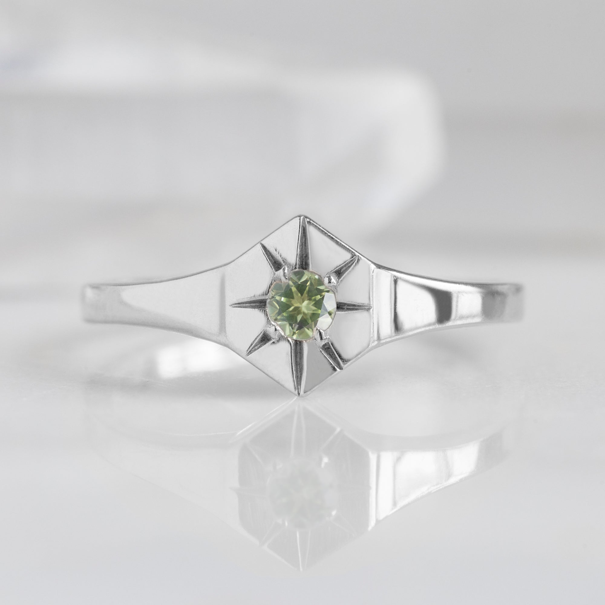 Custom Peridot Birthstone Signet Ring | 10K White Gold. Displayed on marble surface with white blurred background. 