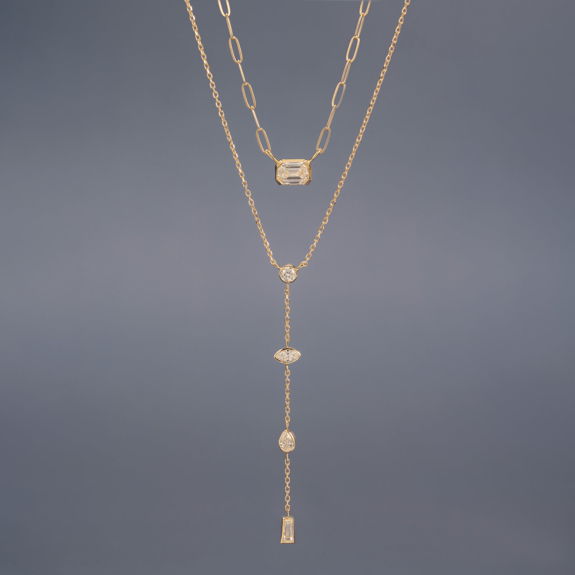 Deco Diamond Solitaire Necklace and Date Night Diamond Lariat Necklace in 10K Yellow Gold hanging in front of muted blue background