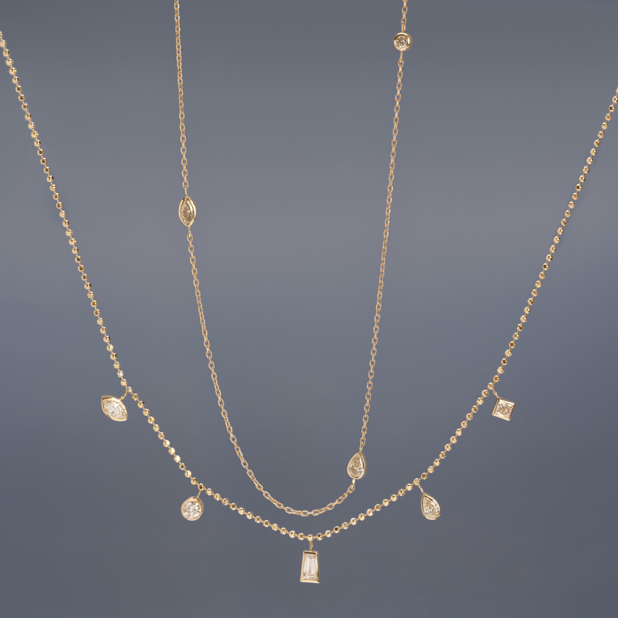 Pear-Cut Diamond Chain and Diamond Pennant Necklaces in 10K Yellow Gold hanging in front of muted blue background