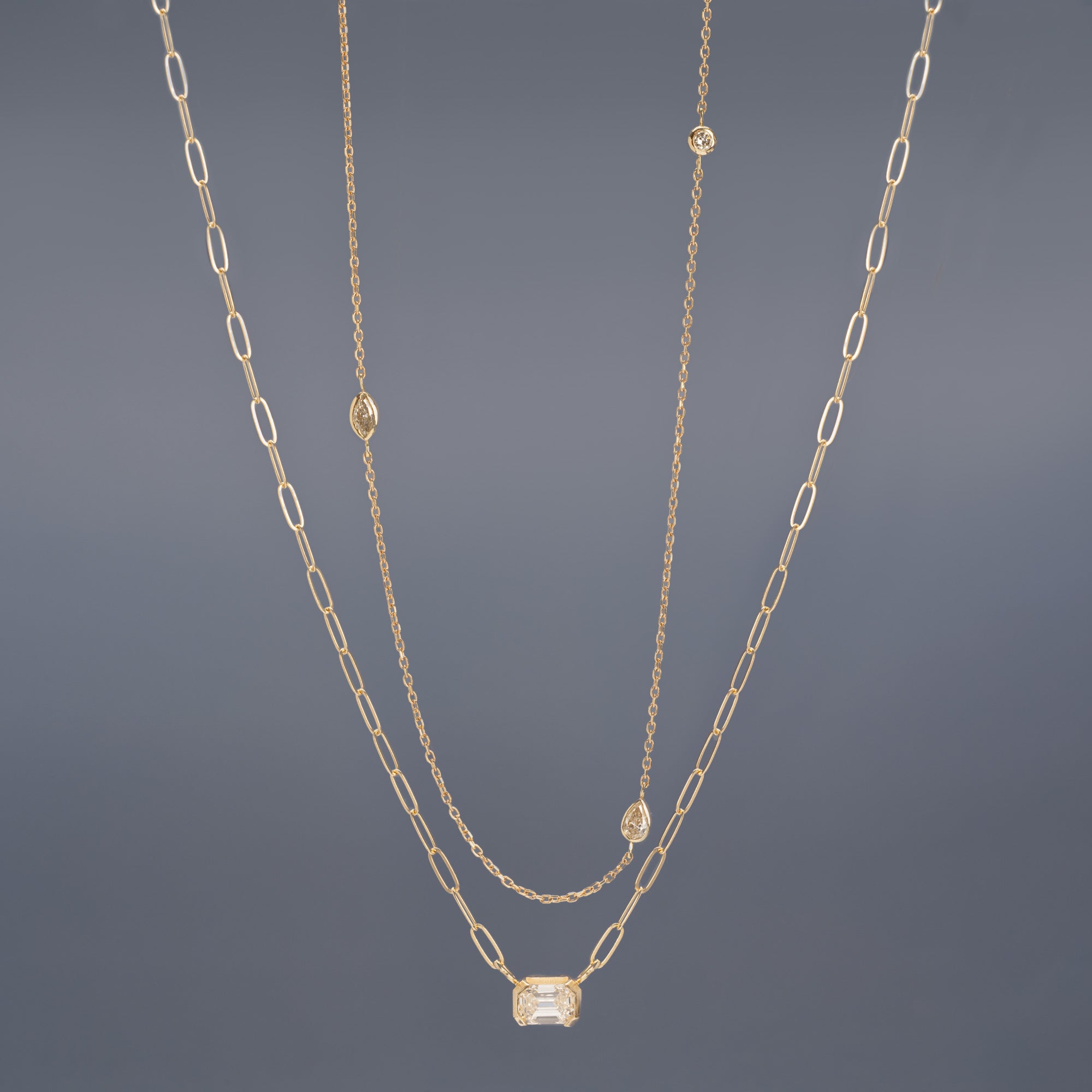 Pear-Cut Diamond Chain and Deco Diamond Solitaire Necklaces in 10K Yellow Gold hanging in front of muted blue background