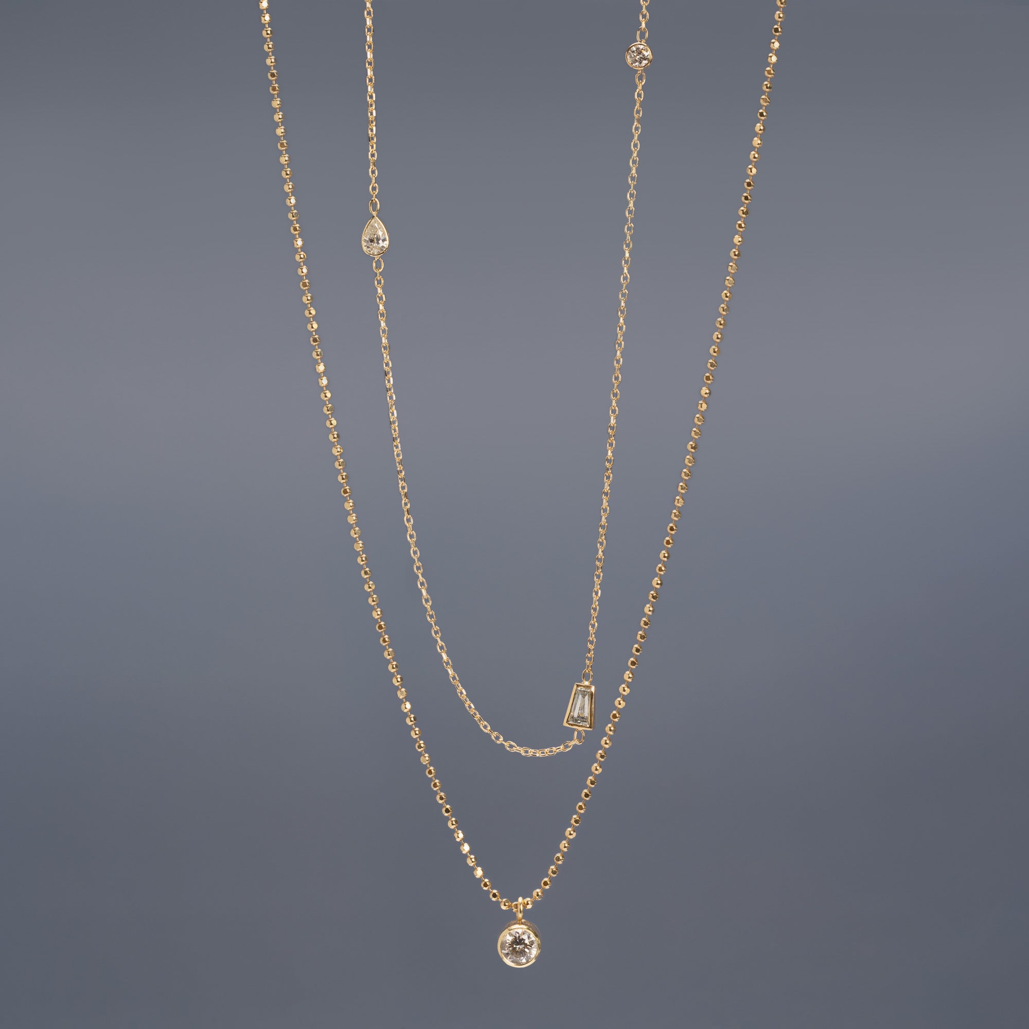 Baguette Diamond Chain Necklace and Not your Typical Solitaire Diamond Necklace in 10K Yellow Gold hanging in front of muted blue background