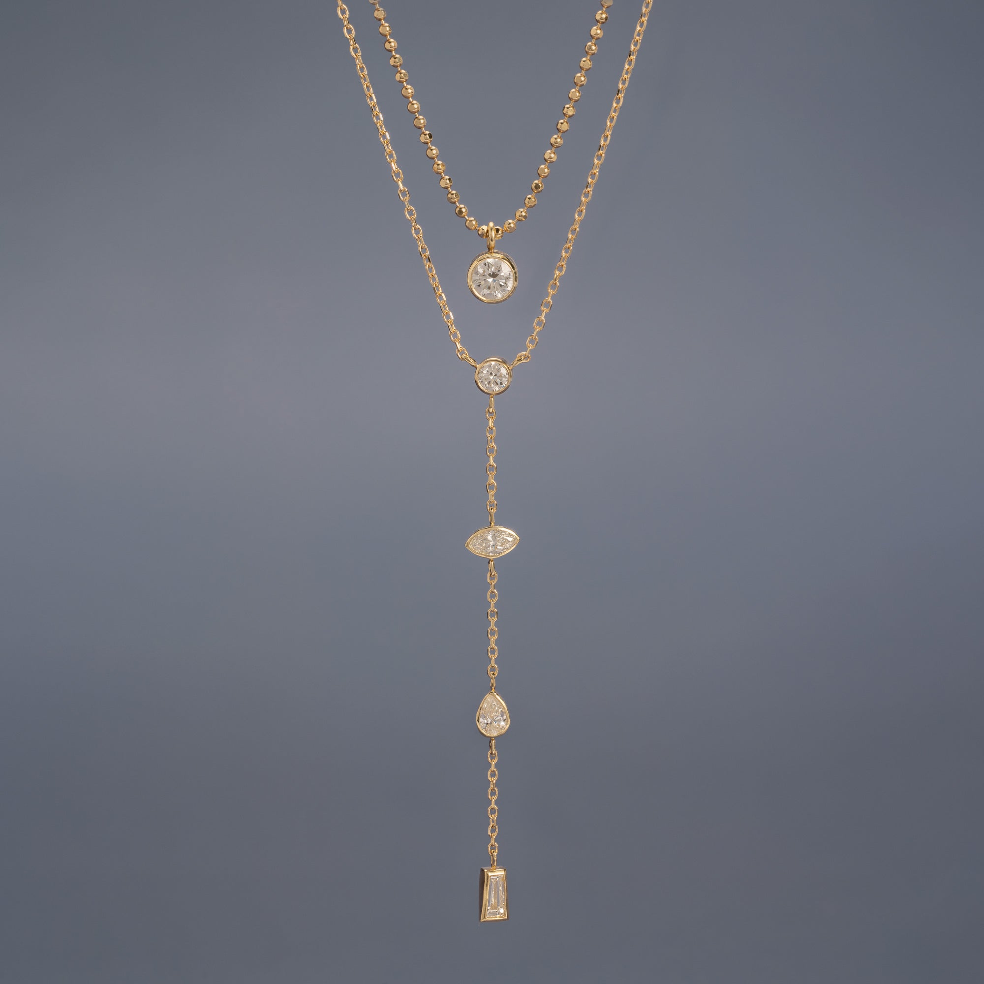 ‘Not Your Typical Solitaire’ Diamond Necklace and 'Date Night' Diamond Lariat 10K Yellow Gold hanging in front of muted blue background