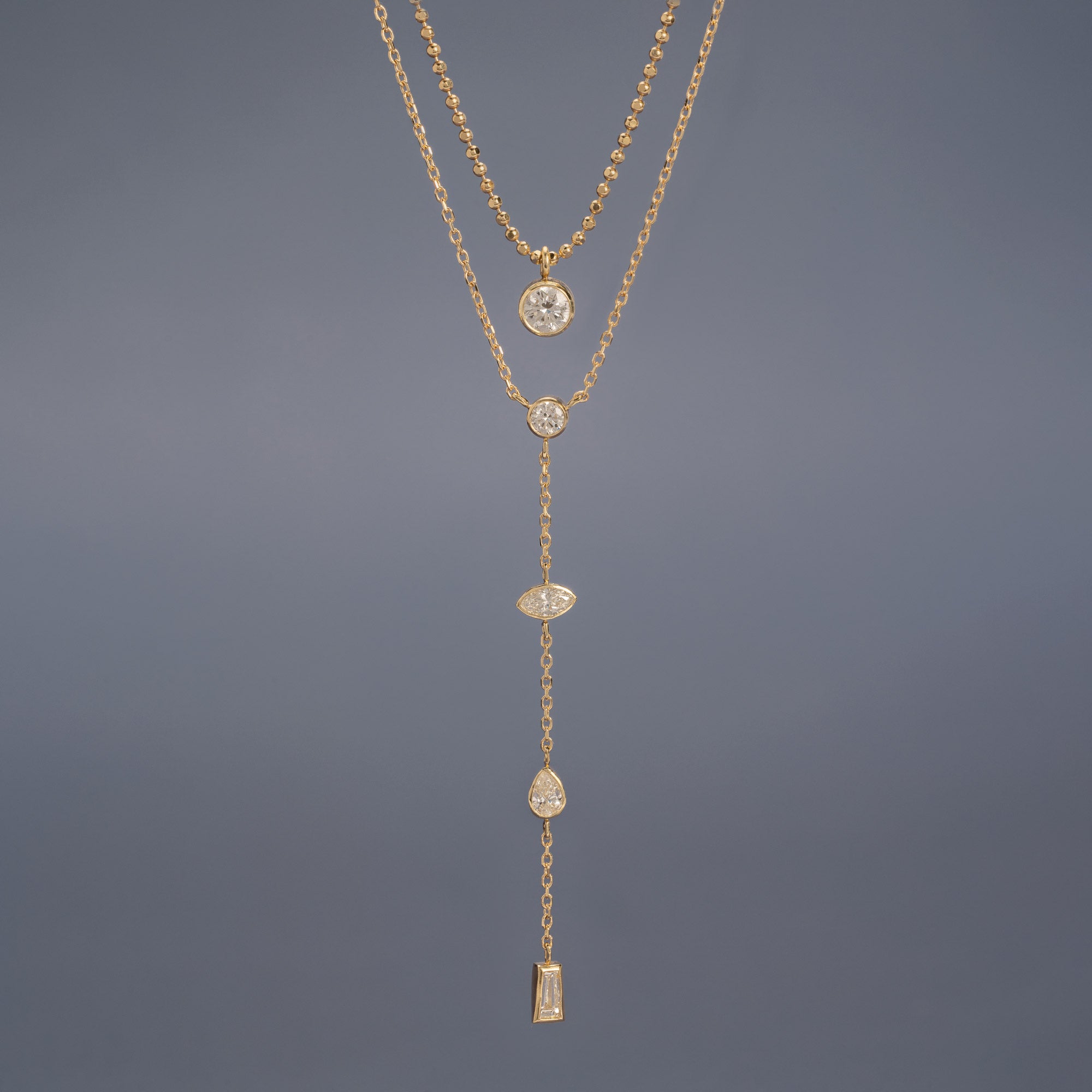 ‘Date Night’ Diamond Lariat Necklace and ‘Not Your Typical Solitaire’ Diamond Necklace in 10K Yellow Gold hanging in front of muted blue background