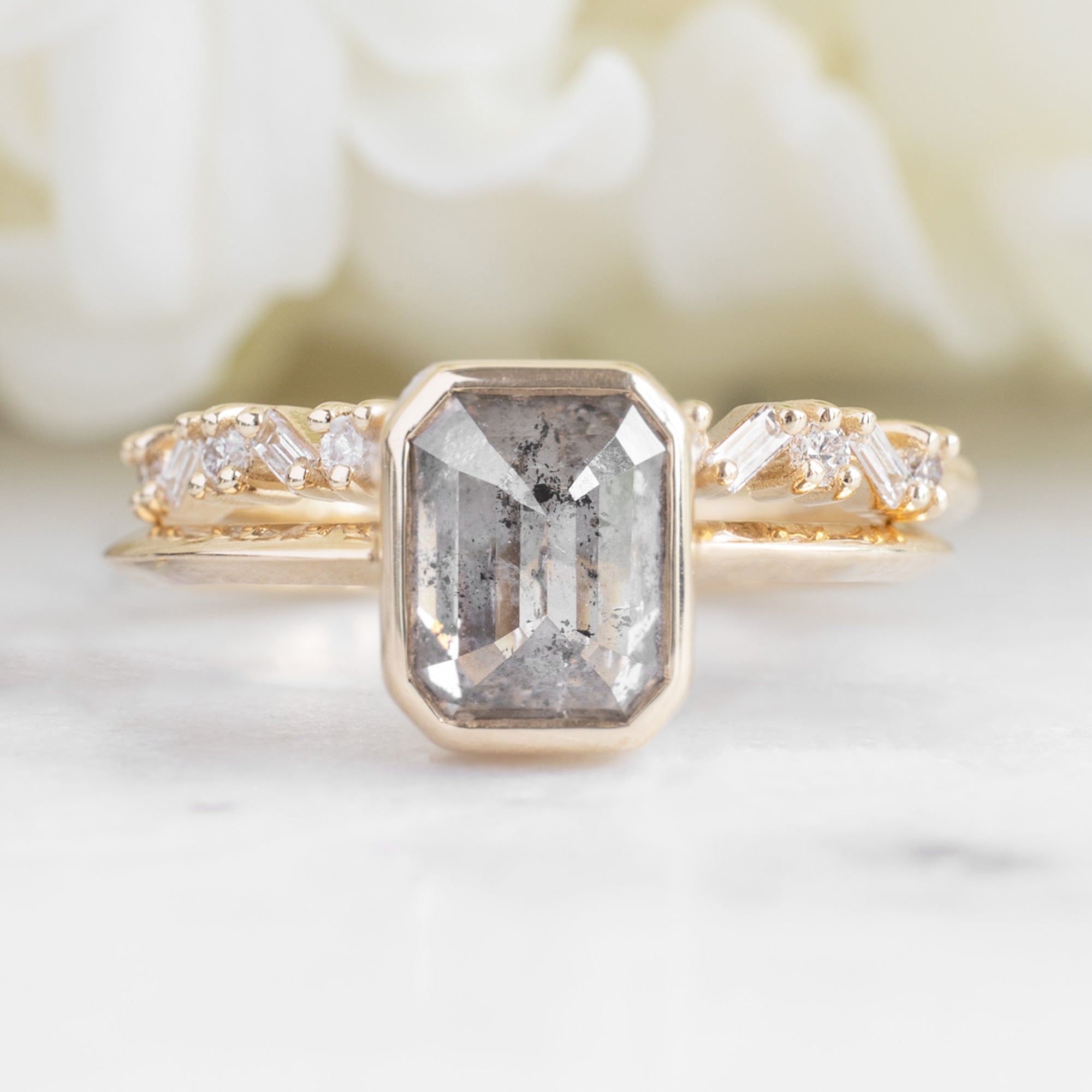 The Baguette Confetti Stacking Ring In Yellow Gold on marble surface paired with an Emerald Cut Bezel Set Diamond Engagement ring.