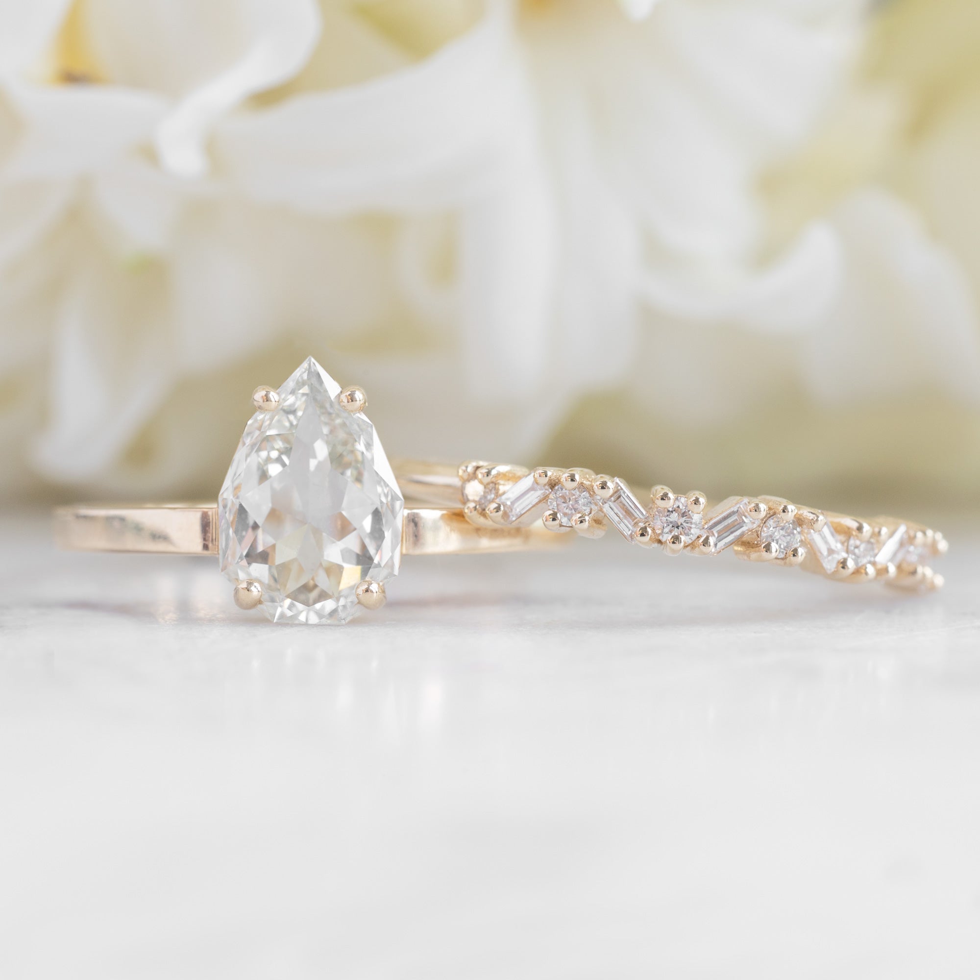 The Baguette Confetti Stacking Ring In Yellow Gold Displayed on a marble surface paired next to a Pear cut Prong Set Diamond Engagement ring.